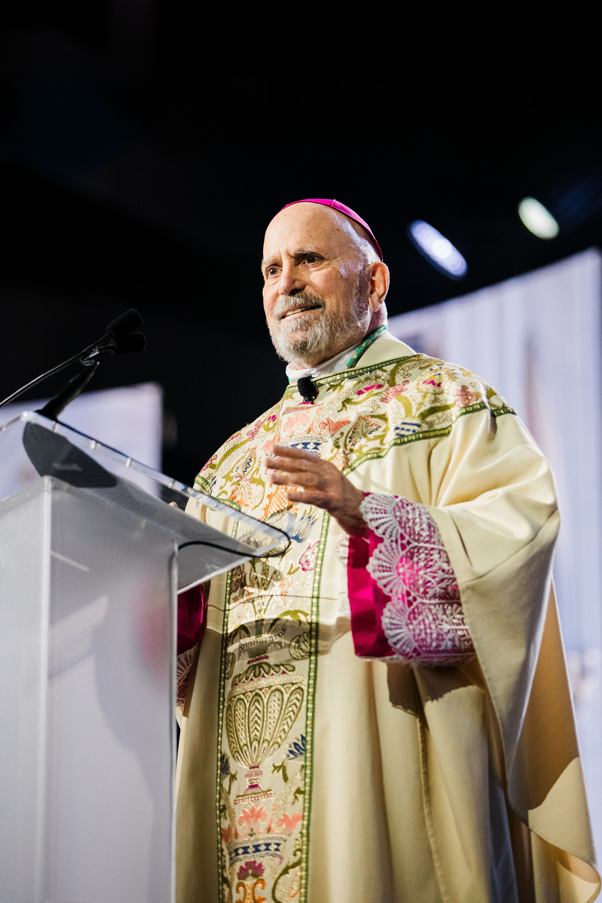 A man in ornate religious robes speaks at a podium. Bright lighting, intricate patterns, and a confident expression create a solemn atmosphere.