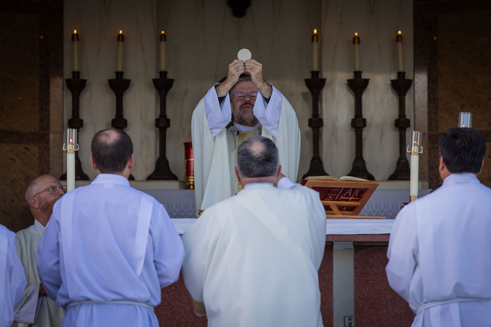 Priest holds up Eucharist during Mass, with altar servers in white robes. Lit candles and open book on the altar. Sacred, solemn setting.
