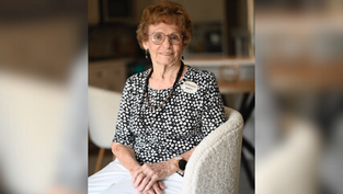Elderly woman with glasses, wearing a black and white polka dot top and a name tag, sits smiling in a cozy room with blurred background.