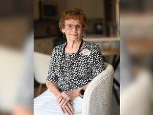 Elderly woman with glasses, wearing a black and white polka dot top and a name tag, sits smiling in a cozy room with blurred background.