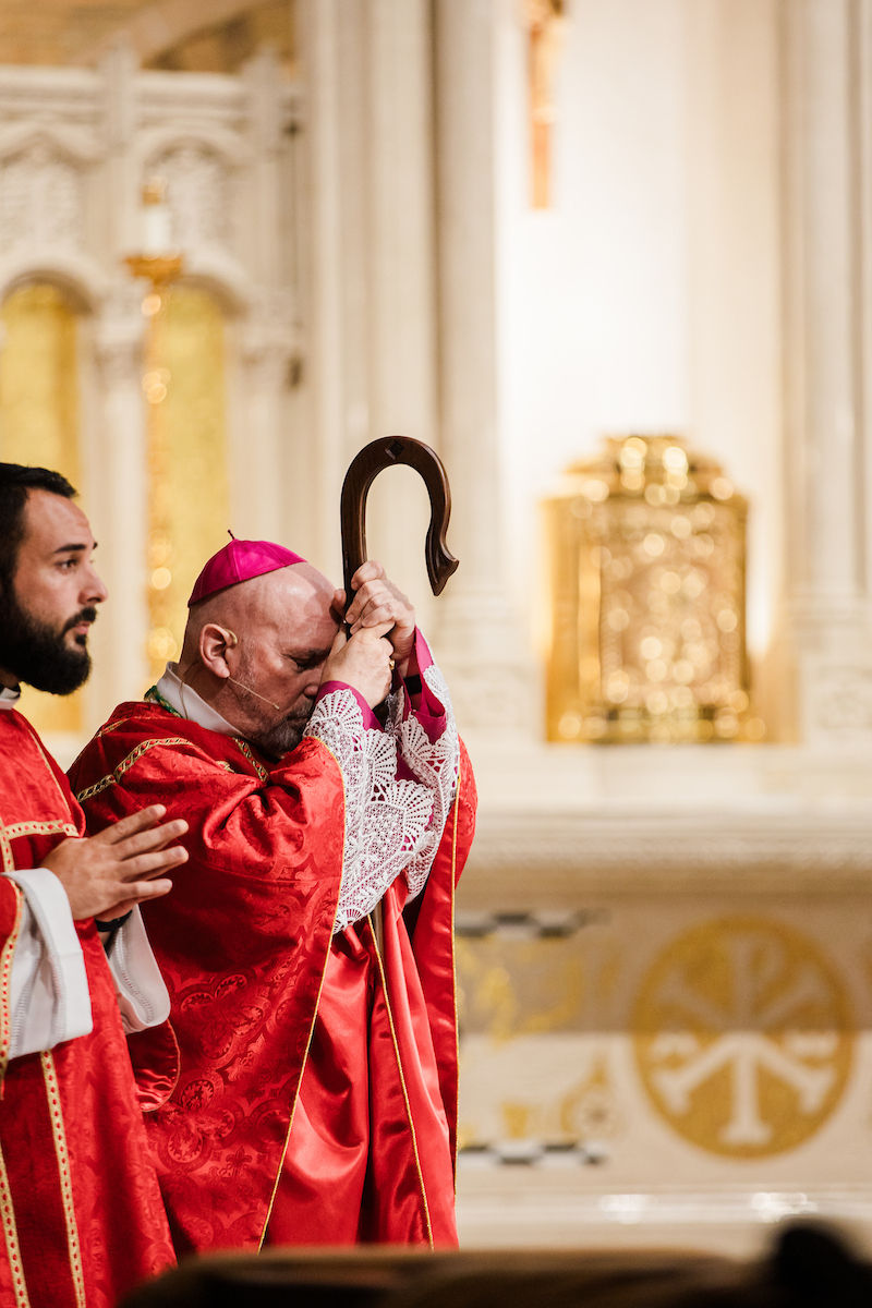 A bishop in a red robe holds a crosier, praying with eyes closed in a church. Another cleric is beside him. Golden ornate background.