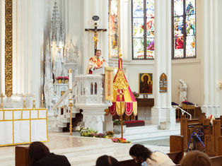 A priest in ornate vestments speaks from a pulpit in a bright church, with stained glass and religious statues in the background.