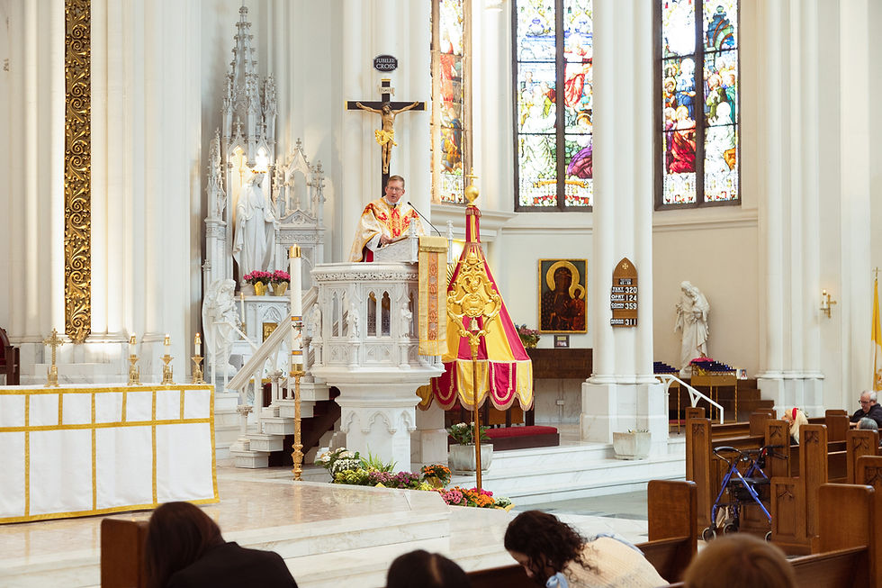 A priest in ornate vestments speaks from a pulpit in a bright church, with stained glass and religious statues in the background.