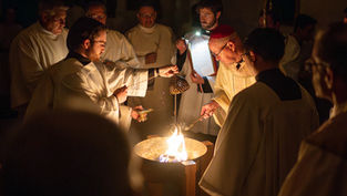 Men in robes perform a religious ritual around a fire inside a church. A candle is lit, and incense is swung, creating a solemn atmosphere.