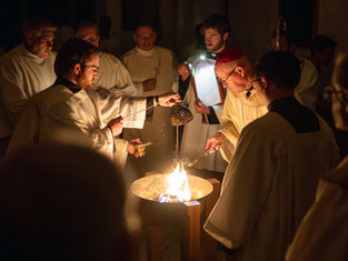 Men in robes perform a religious ritual around a fire inside a church. A candle is lit, and incense is swung, creating a solemn atmosphere.