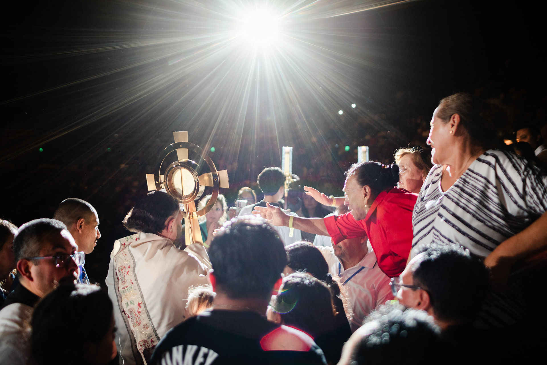 A crowd gathers around a person holding a golden religious object under a bright light. People reach out, wearing colorful clothing.