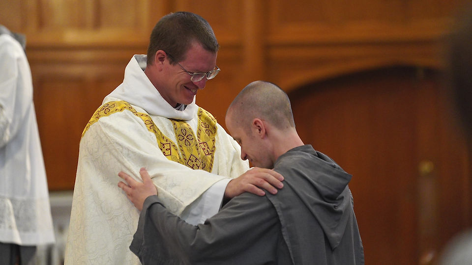 Priest in white and gold robes smiling warmly, blesses a kneeling man in gray in a wooden church setting.