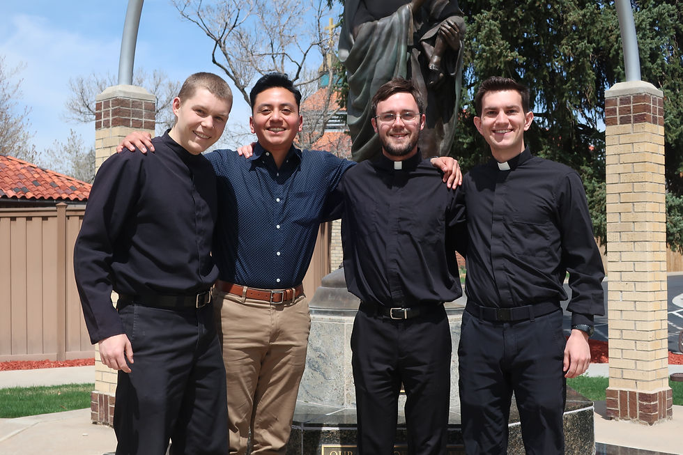 Four men stand smiling, arms around each other, in front of a statue outdoors. Two wear clerical shirts, the other two wear casual shirts.