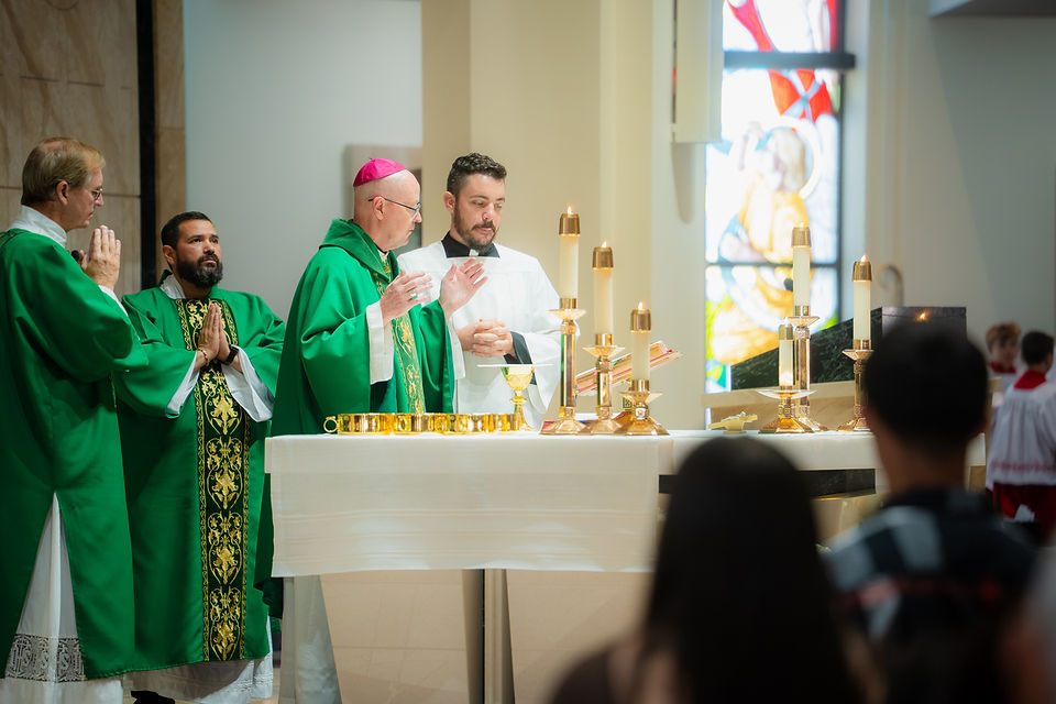 Catholic priests in green vestments pray at a candlelit altar in a church, with stained glass and worshippers blurred in front.