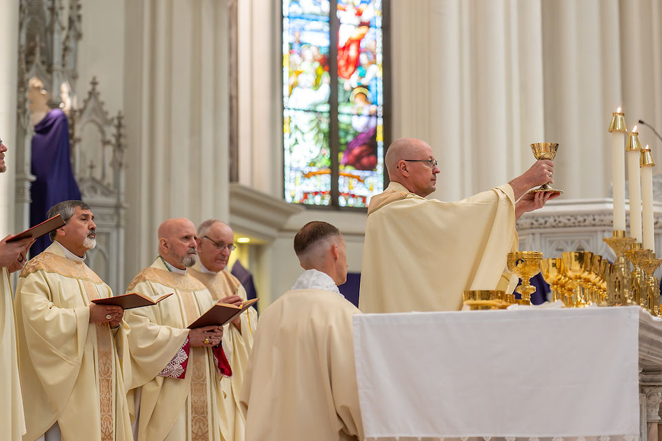Clergy in beige robes perform a Eucharistic rite in a church, with one priest raising a chalice. Stained glass window in the background.