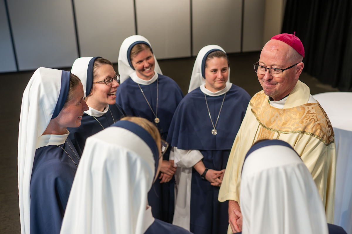 A bishop in ornate robes converses with a group of nuns in navy habits, smiling in a neutral-colored room. The mood is friendly and warm.