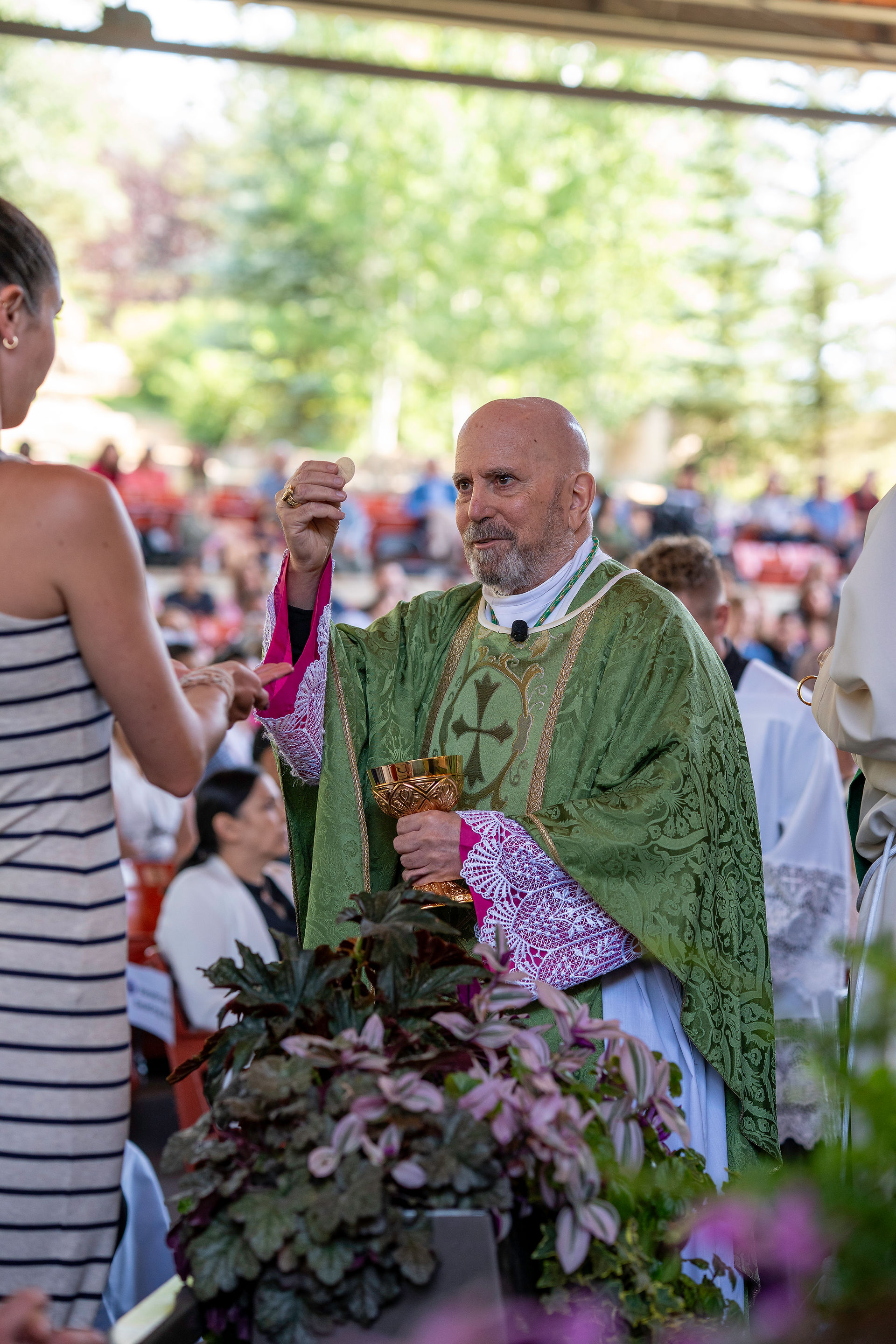 A priest in a green and gold chasuble administers Holy Communion to a congregant during an outdoor Catholic Mass. He holds a chalice and offers a communion wafer to a woman in a striped dress, who receives it with an outstretched hand. Other attendees and greenery are visible in the background.