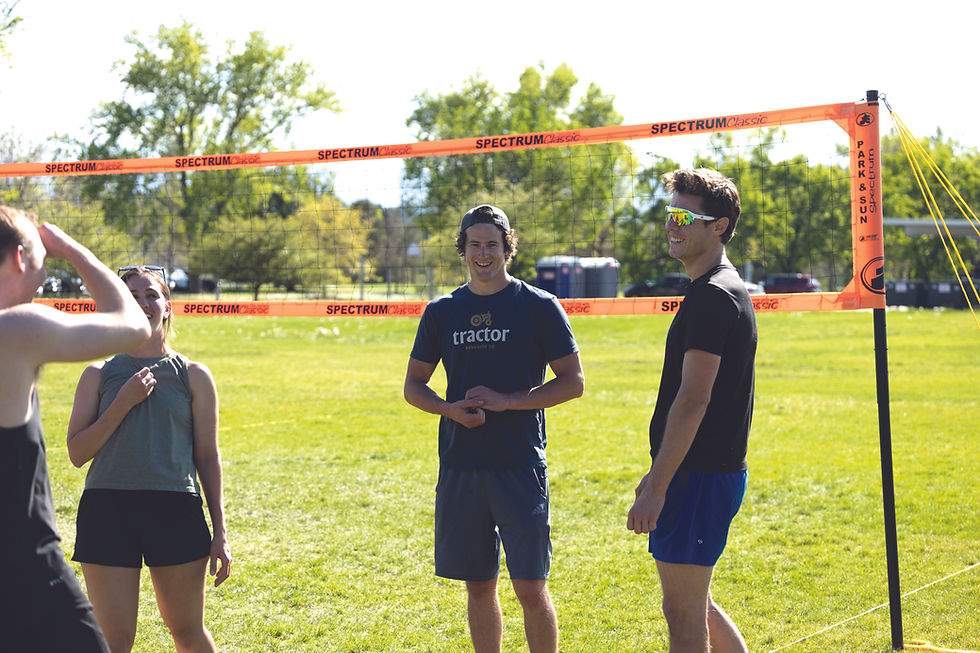 Four people in athletic clothing stand in front of a volleyball net, casually conversing and smiling. The grass and trees are green, giving a warm feeling. The mood is light and fun.