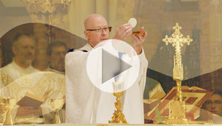 A bishop in white vestments holds the Eucharist up during Mass, against a stylized background with various swoosh shapes at different levels of contrast. A play button is superimposed, indicating that this is a featured video image. The mood is prayerful.
