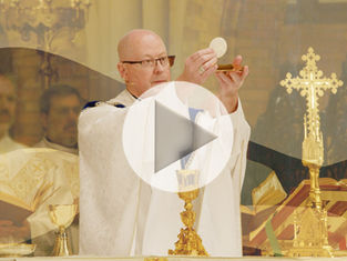 A bishop in white vestments holds the Eucharist up during Mass, against a stylized background with various swoosh shapes at different levels of contrast. A play button is superimposed, indicating that this is a featured video image. The mood is prayerful.