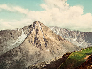 Snow-streaked mountain peaks under large white clouds and a pale blue sky, with green and rocky foreground.