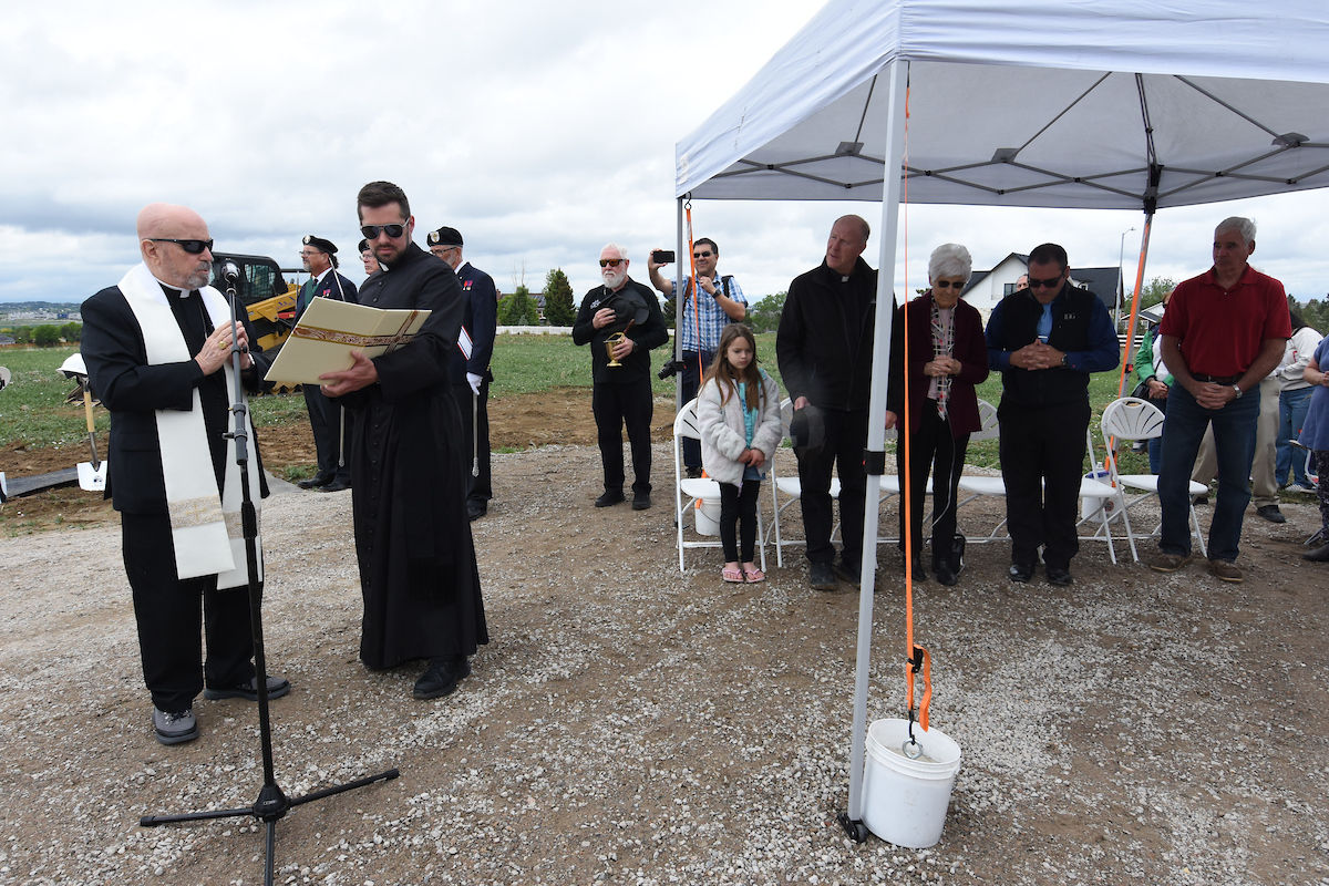 A priest reads from a book at a ceremony under a cloudy sky, with attendees standing and sitting under a white tent on gravel.