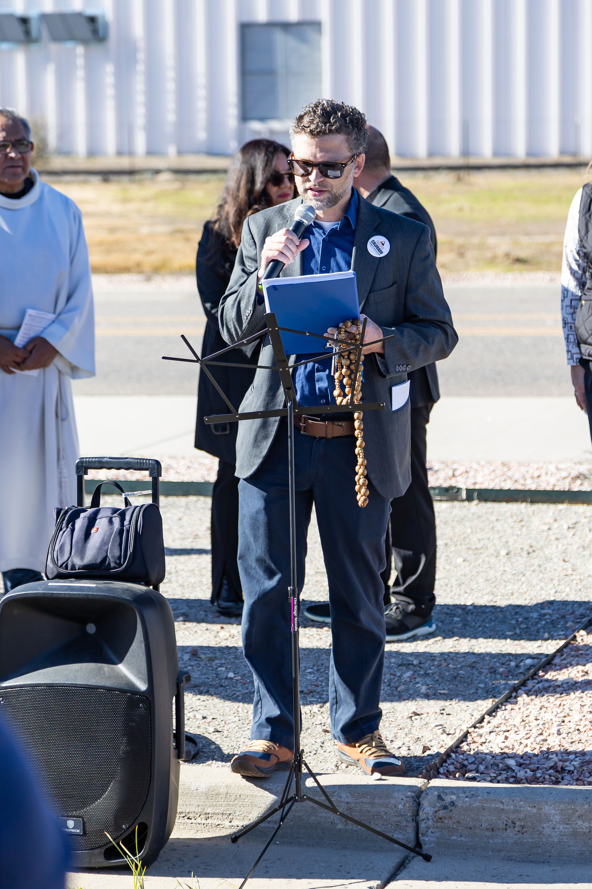 Man in a suit speaks into a microphone, holding notes and rosary, outside on a sunny day. Speaker and luggage in the background. Casual setting.