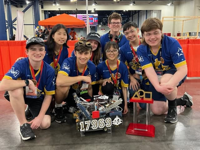 High school robotics team students in a convention center wearing blue jerseys with their robot and an award they won in front of them.