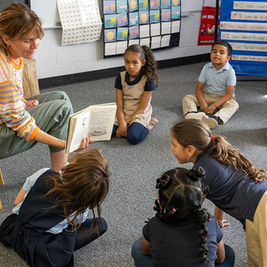 Teacher reading to attentive children sitting on a classroom floor. Bright, colorful setting with educational posters on walls.