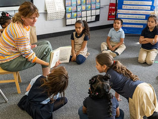 Teacher reading to attentive children sitting on a classroom floor. Bright, colorful setting with educational posters on walls.