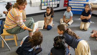 Teacher reading to attentive children sitting on a classroom floor. Bright, colorful setting with educational posters on walls.
