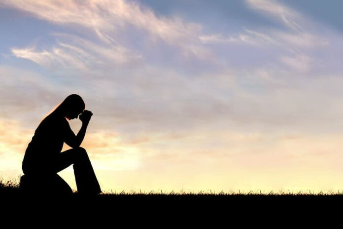 A silhouette of a young Christian woman is bowing her head in prayer, and desperation outside during sunset.