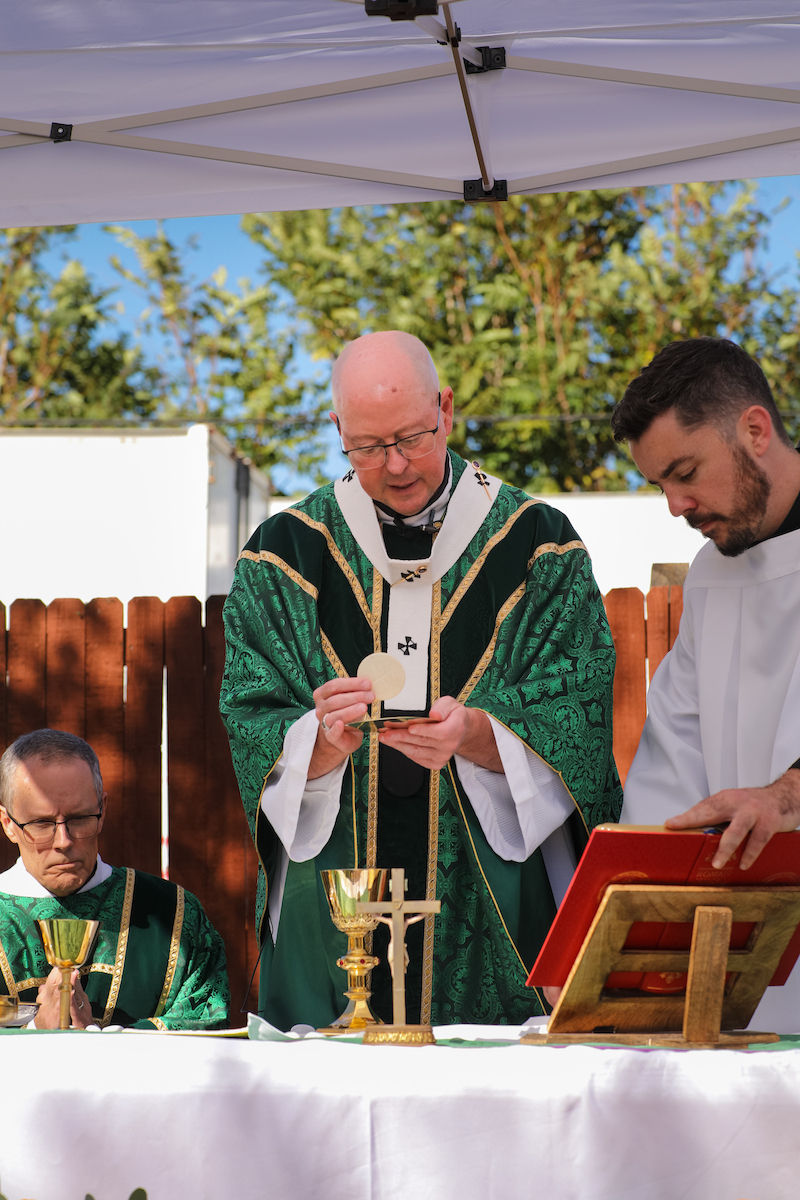 Priests in green vestments celebrate outdoor Catholic Mass under a white canopy, one lifting the Eucharist at the altar.