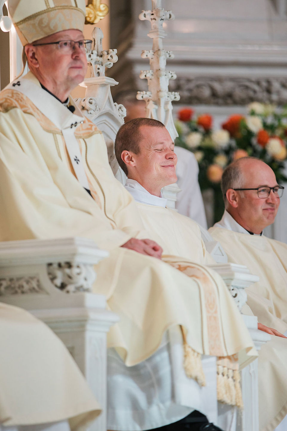 Three Catholic clergy in cream vestments sit during a ceremony, one smiling with eyes closed, in an ornate church.