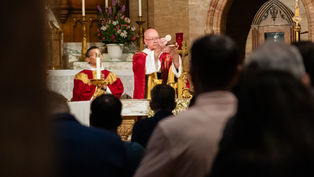 Archbishop elevates communion wafer at altar during Catholic Mass, with candlelit church interior and worshippers in foreground.