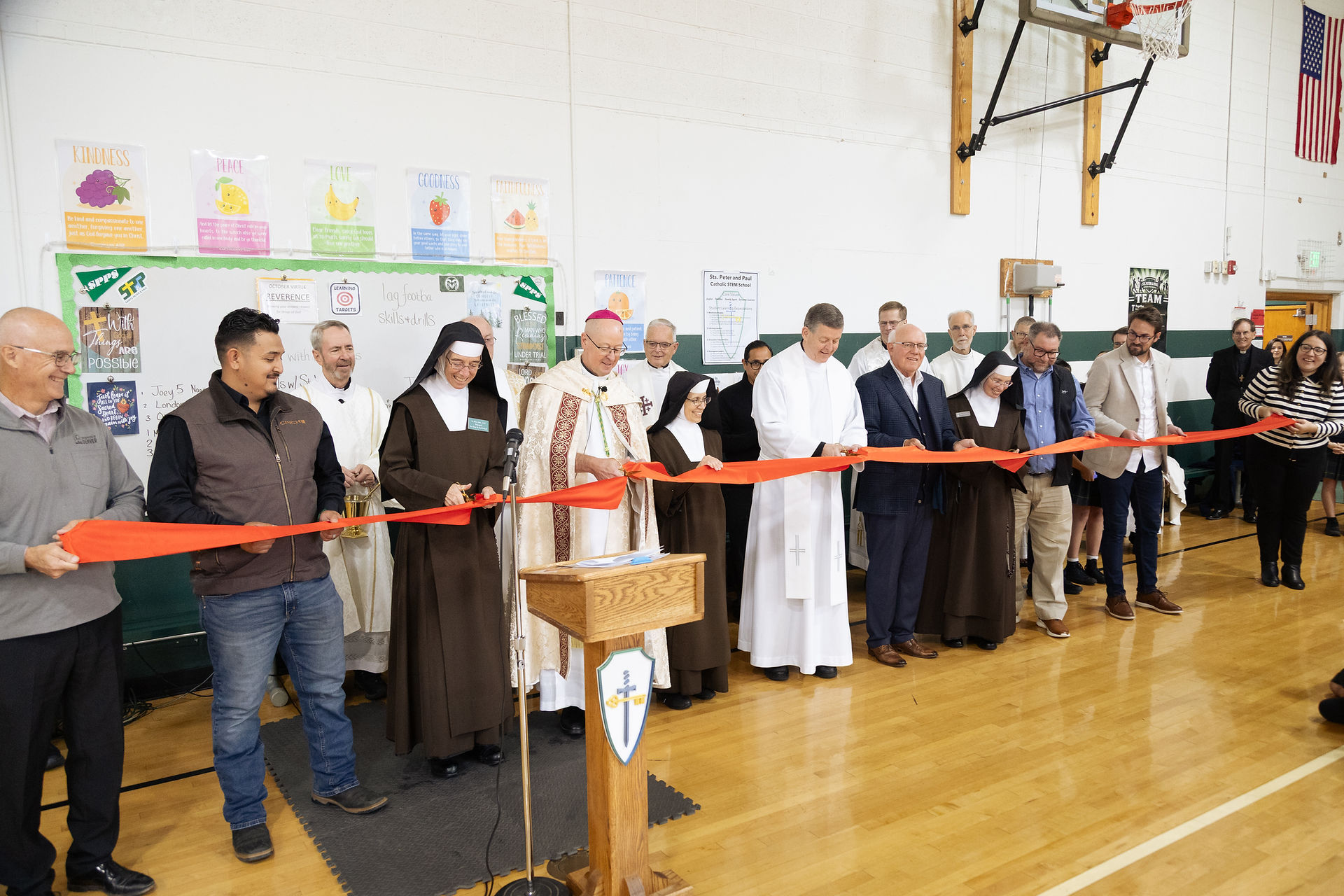 Ribbon-cutting ceremony in a school gym, with clergy and adults holding a red ribbon beneath virtue posters.