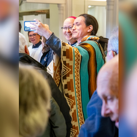Man in a patterned turquoise poncho takes a selfie with smiling people in a crowded indoor hall, phone raised high.