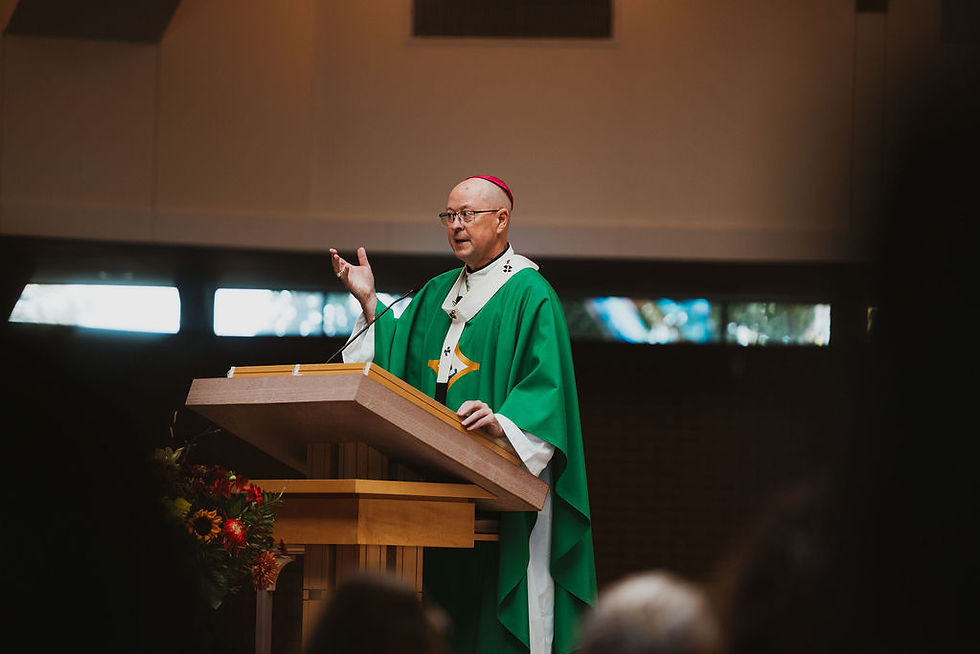 Bishop in green vestments speaks at a church lectern, one hand raised, with a floral arrangement and blurred audience below.