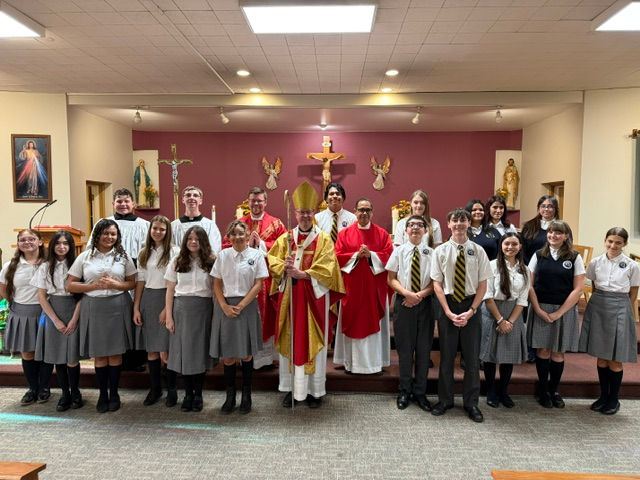 Students and clergy pose on a church altar, with a bishop in gold and red centered before crucifix and statues.