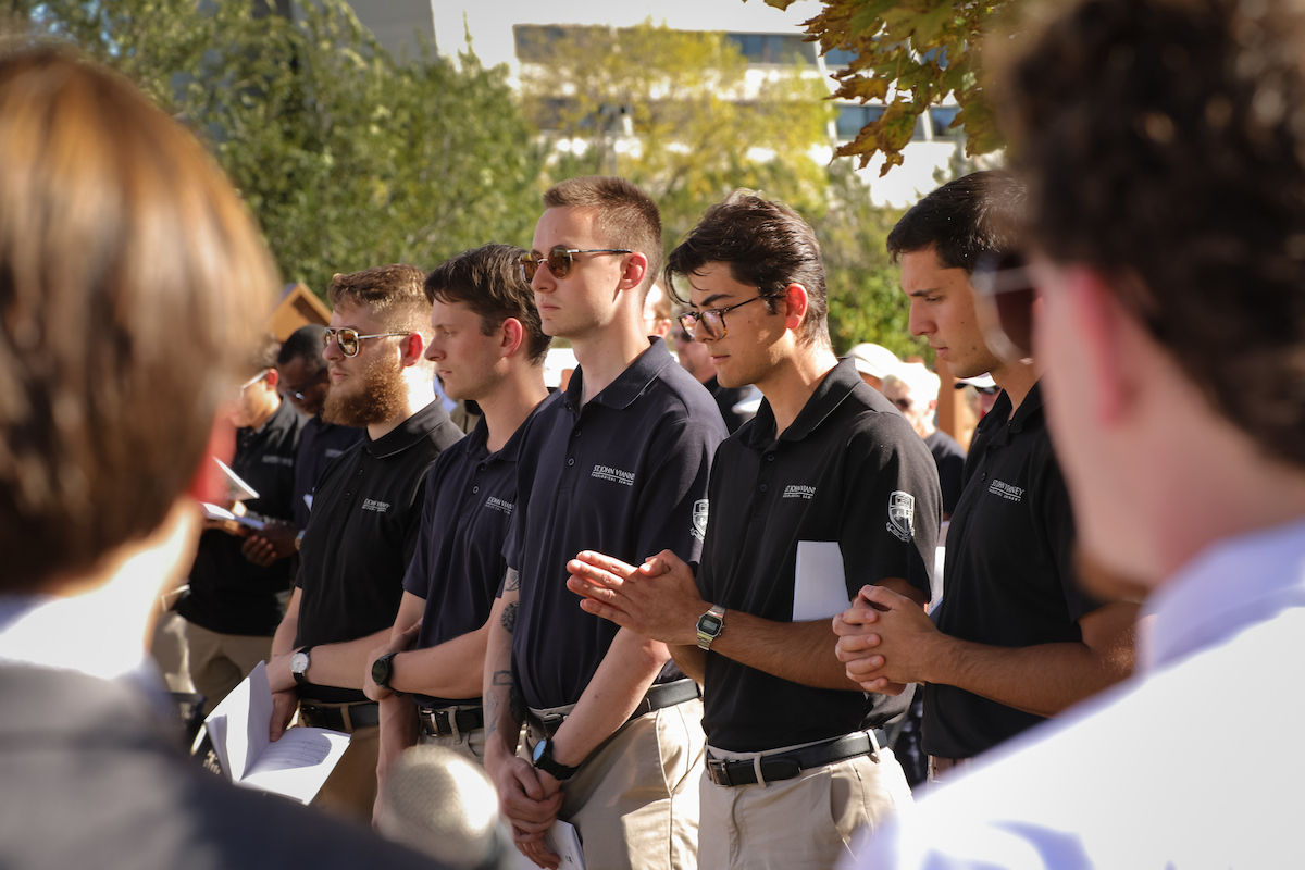 Young men in navy polos and khakis stand outdoors on campus in a solemn gathering, some clasping hands and holding papers.