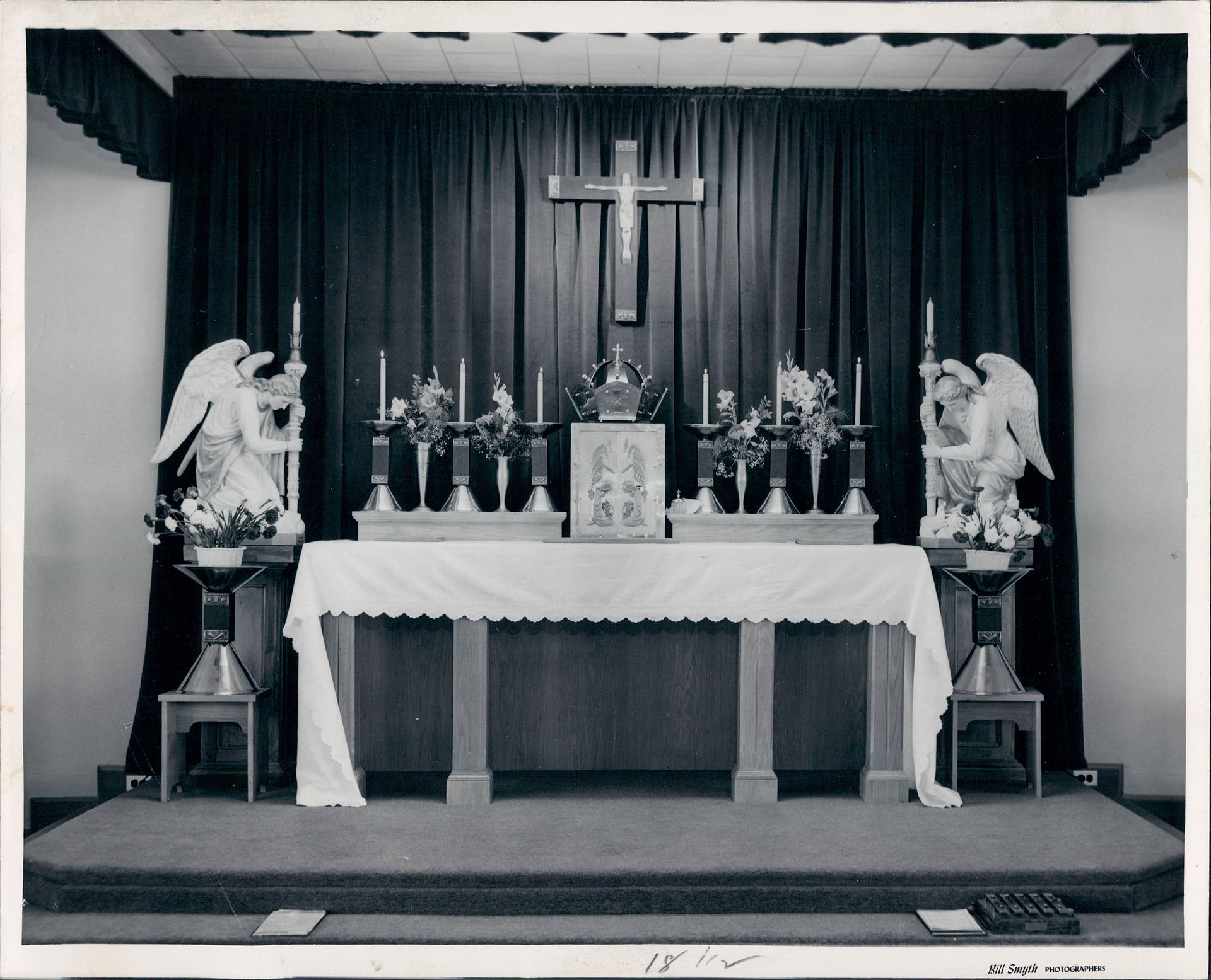 Black-and-white church altar with crucifix, candles, flowers, and kneeling angel statues before dark curtains