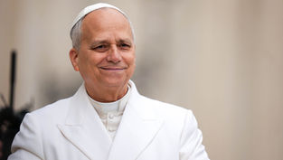 Smiling pope in white papal robes and skullcap against a soft beige background, looking calm and warm