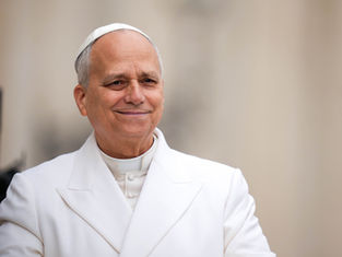 Smiling pope in white papal robes and skullcap against a soft beige background, looking calm and warm