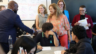 Adults shake hands in a classroom while students sit at desks; classroom charter poster and TV screen in background.