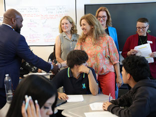 Adults shake hands in a classroom while students sit at desks; classroom charter poster and TV screen in background.