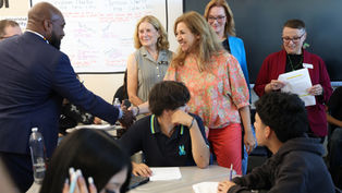Adults shake hands in a classroom while students sit at desks; classroom charter poster and TV screen in background.