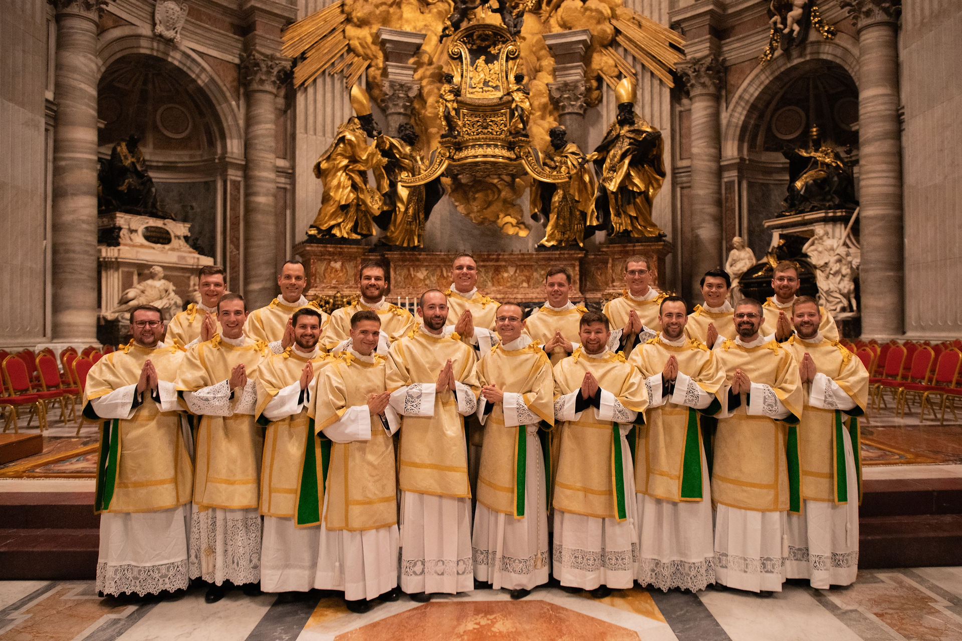 Group of deacons in gold vestments pose smiling before a grand gilded altar in an ornate cathedral.