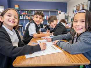 Smiling elementary students at a classroom table with worksheets and markers, learning together in a cheerful classroom.