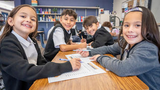 Smiling elementary students at a classroom table with worksheets and markers, learning together in a cheerful classroom.