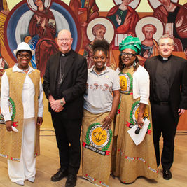 Five smiling people, including two priests, pose before a colorful saint mural; African Catholic Society garments and hats stand out.