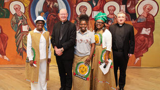 Five smiling people, including two priests, pose before a colorful saint mural; African Catholic Society garments and hats stand out.