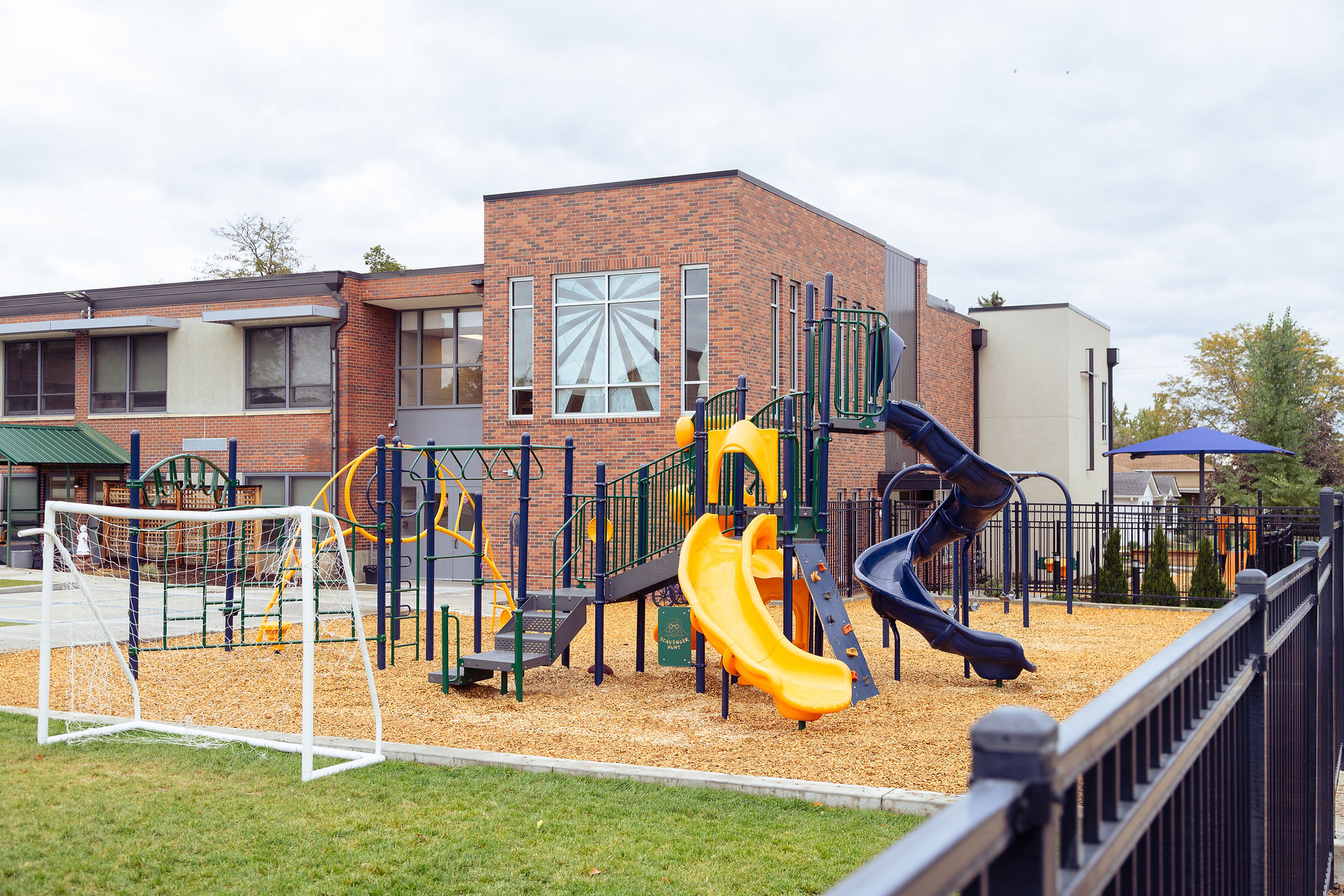 Empty playground with yellow and navy slides beside a brick building; small green sign reads scavenger hunt.