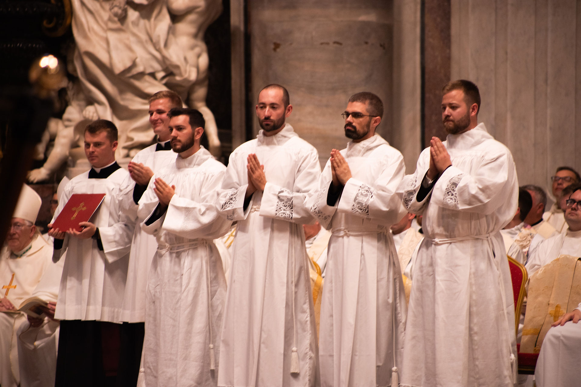 Five men in white robes stand praying in a church, one holding a red book with a gold cross, with clergy seated behind them.