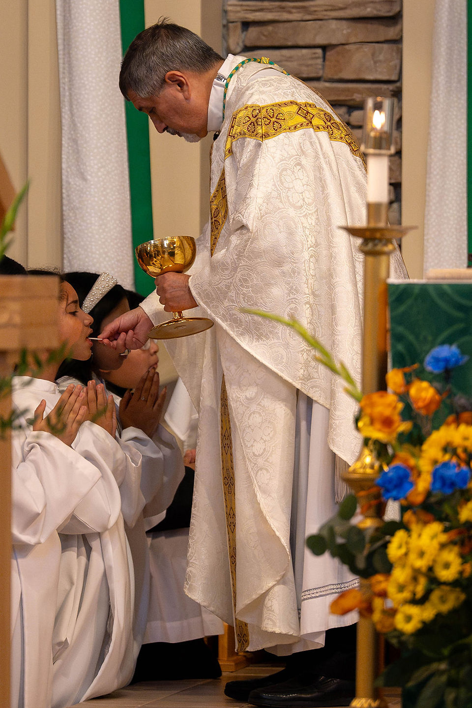 Priest in white-and-gold vestments gives communion to kneeling children during a church service, with flowers and candles nearby.