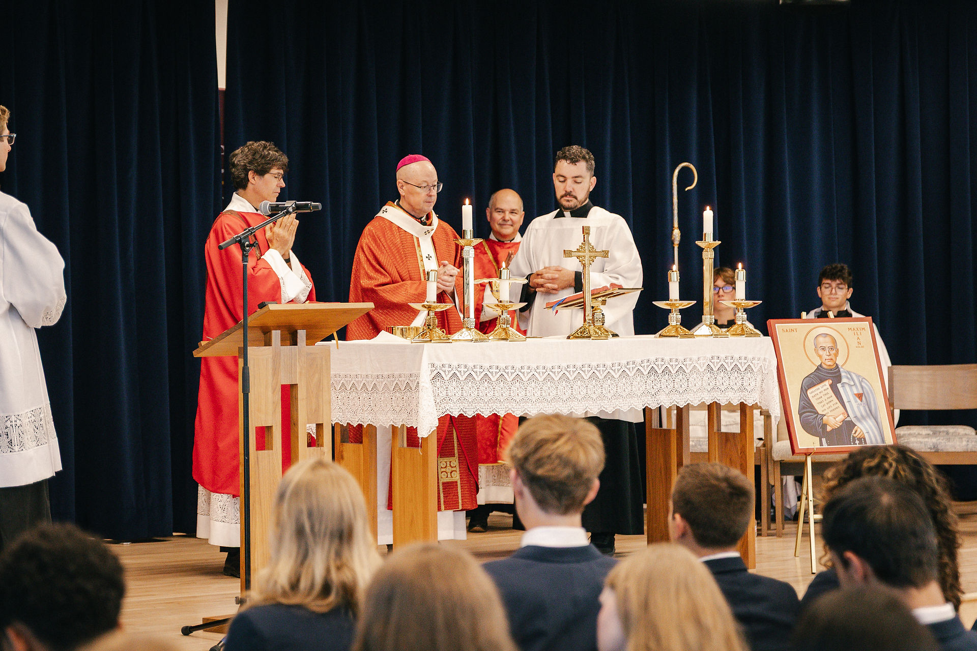 Clergy in red and white vestments lead a candlelit church service before an altar, with a Saint Maximilian icon and seated congregation.